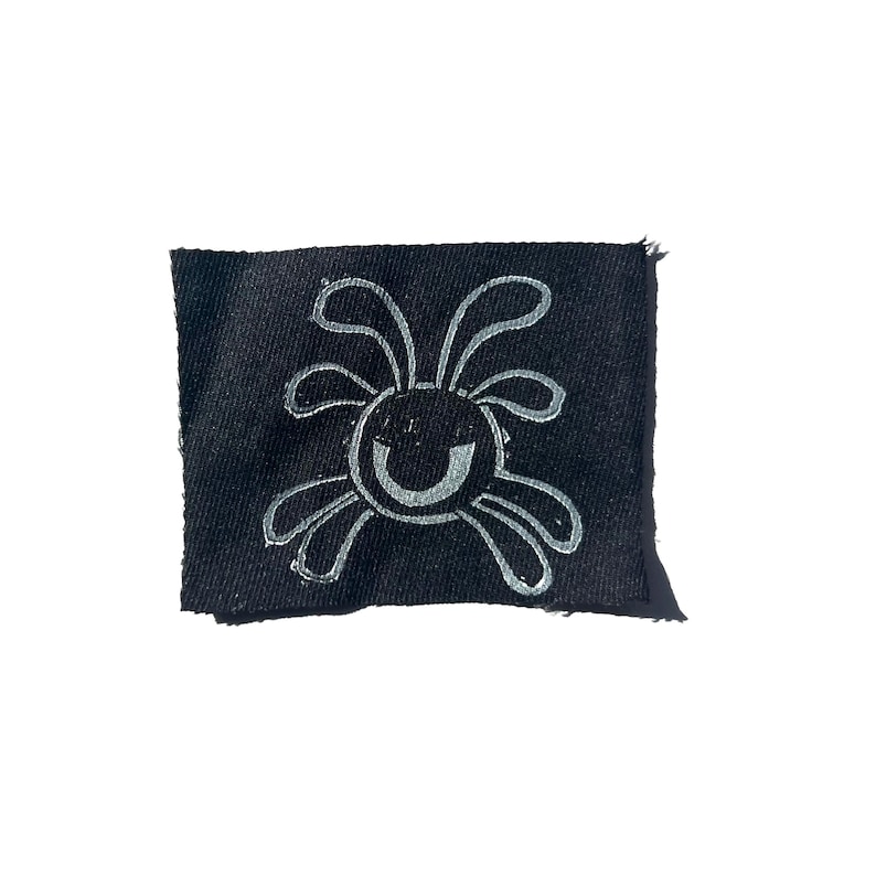 May include: A small, black fabric patch featuring a whimsical, smiling sun design in silver. The sun has curved lines for rays and a curved line for the mouth. The patch has a raw edge.