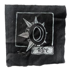 May include: Black fabric patch with a silver compass rose design inside a square frame. The word "Rare" is printed on a small rectangular label below the compass rose.