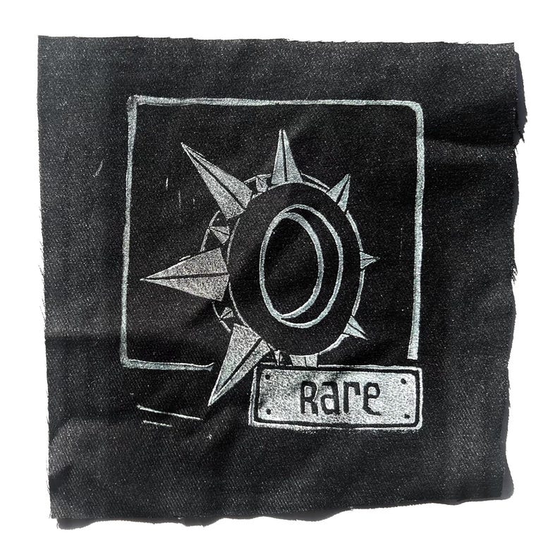 May include: Black fabric patch with a silver compass rose design inside a square frame. The word "Rare" is printed on a small rectangular label below the compass rose.