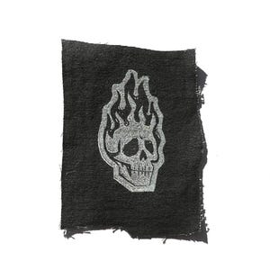 May include: A black fabric patch with a silver skull design engulfed in flames. The edges of the patch are frayed, giving it a distressed look. The skull and flames are detailed with shading, creating a three-dimensional effect.