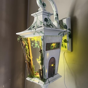 May include: A white wall-mounted lantern featuring hand-painted green vines and leaves. The interior contains a miniature scene with a small fountain, moss, and twigs. The lantern has a rectangular window and an arched door, illuminated by a warm light.