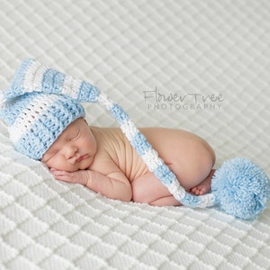 May include: A newborn baby wearing a blue and white striped crocheted hat with a pom-pom on the end. The baby is sleeping on a white blanket with a diamond pattern.