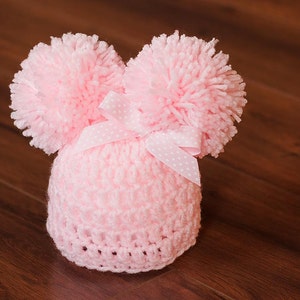 Puede incluir: Un gorro de beb&eacute; de punto rosa con dos pompones esponjosos en la parte superior y un lazo blanco con lunares.