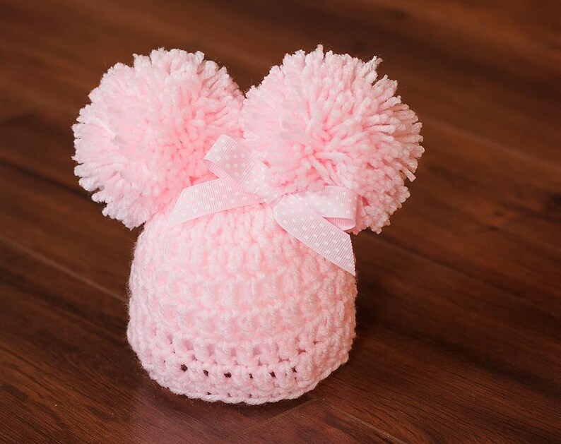 Puede incluir: Un gorro de beb&eacute; de punto rosa con dos pompones esponjosos en la parte superior y un lazo blanco con lunares.