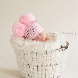 Puede incluir: Un gorro de beb&eacute; de crochet rosa con dos grandes pompones en la parte superior. El gorro lo lleva puesto un beb&eacute; durmiendo en una cesta blanca y esponjosa.