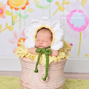 May include: A newborn baby wearing a white crocheted daisy flower hat with green ribbon, sleeping in a light brown flower pot on a green rug. The background is a watercolor painting of flowers.