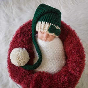 May include: A newborn baby wrapped in a white blanket, wearing a green and cream-colored crochet hat with a bow and a long tail. The baby is nestled in a red, fluffy nest with a cream-colored pom-pom.