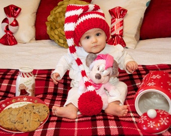 May include: A baby wearing a red and white striped knit hat with a long tassel, sitting on a red plaid blanket. A plush toy, cookies, and a milk bottle are nearby, creating a festive holiday scene.