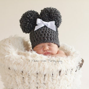 May include: A newborn baby wearing a gray crocheted hat with two pom-poms and a white bow, sleeping in a white fluffy basket.
