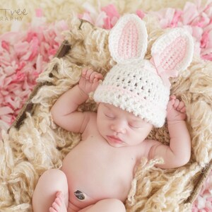 May include: A newborn baby wearing a white crocheted bunny hat with pink ears, sleeping in a basket filled with light brown fluffy material. The basket is on a pink and white background.