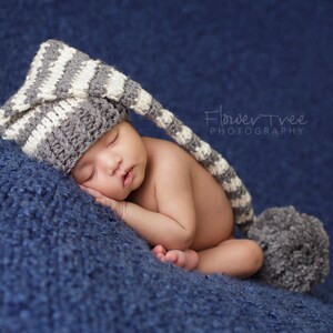 May include: A baby wearing a gray and white striped knitted hat with a long tail sleeps on a blue fuzzy blanket. The hat has a long tail that is tucked under the baby's body.