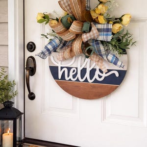 May include: A circular wooden welcome sign with the word "hello" in white script. The sign is adorned with yellow flowers, green leaves, and plaid ribbons in blue, brown, and beige. The sign is suspended on a white door.