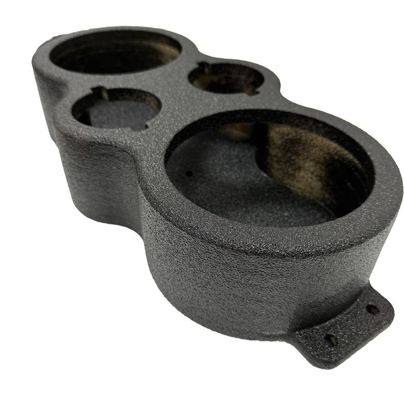 May include: A textured, dark gray automotive gauge pod with multiple circular openings of varying sizes. The pod has a curved shape and a mounting bracket with two holes.