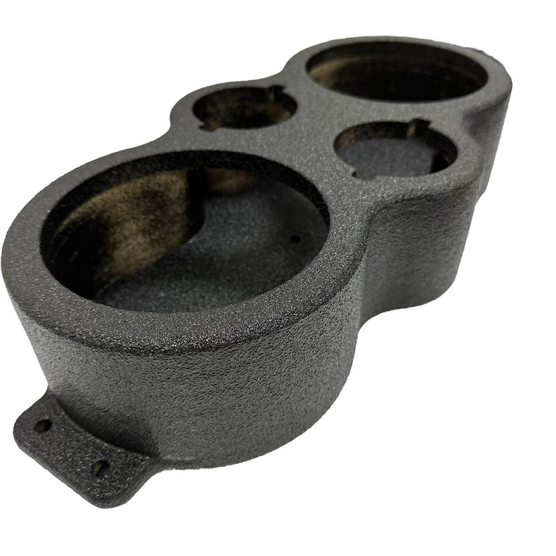 May include: A textured, dark gray automotive gauge pod with three circular openings of varying sizes. The pod has a curved shape with a mounting bracket featuring two holes. The surface has a slightly rough, speckled appearance.