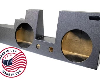 May include: A dark gray speaker box with two large circular openings and several smaller holes. The box has a textured surface and a rectangular shape. The image includes a "Made in USA" sticker with the American flag.