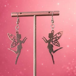 May include: A pair of silver-toned metal earrings featuring a cutout silhouette of a fairy with detailed filigree wings. The earrings hang from standard silver-toned metal fishhook wires, suspended from a horizontal metal display bar.