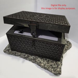 May include: A dark grey wooden box with a decorative lattice design on the lid and sides. The lid is slightly open, revealing a white interior with a cannabis leaf design. The box is secured with a latch and sits on a grey stone. Text on the image reads: "Digital file only. this image is for display purposes."