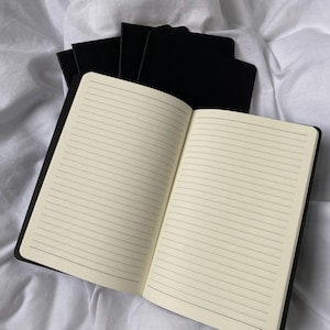 Puede incluir: Varios cuadernos negros con páginas rayadas. Uno de ellos está abierto, mostrando páginas color crema con líneas horizontales. Los cuadernos están colocados sobre una superficie blanca y texturizada.