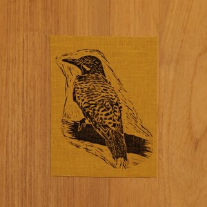 May include: A black and white linocut print of a bird perched on a branch, set against a mustard yellow background. The bird has detailed feather patterns. The print is on a textured fabric.