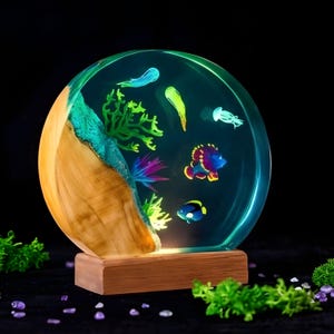 May include: A round, illuminated resin lamp featuring an underwater scene. The lamp has a wooden base and a clear resin globe with colorful fish, coral, and jellyfish. The wood grain is visible on the side.