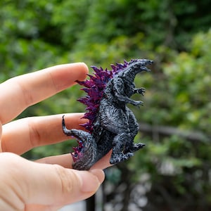 May include: A miniature Godzilla figurine, predominantly dark gray with vibrant purple dorsal fins. The detailed sculpture captures the iconic monster's form, showcasing its textured skin and menacing posture. The figurine is held against a blurred green background.