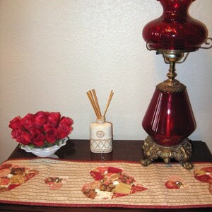 May include: A wooden table with a red and brown patchwork table runner. A red glass lamp with a gold base sits on the table. A small white bottle with a gold top and wooden sticks sits on the table. A bowl of red roses sits on the table.