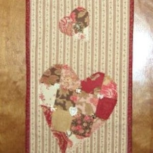 以下が含まれることがあります： 茶色と白のストライプの布製の壁掛けで、ハートのデザインが施されています。ハートはピンク、赤、茶色のパッチワーク生地で作られています。ハートは小さなハートで囲まれています。