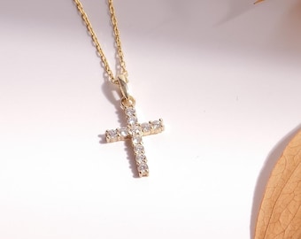 May include: A gold-colored necklace with a cross pendant. The cross is encrusted with small, clear gemstones. The necklace is displayed on a white surface with dried leaves and a wooden element in the background.