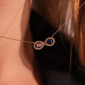 May include: A delicate gold necklace featuring an infinity symbol pendant. The pendant is adorned with small, sparkling crystals and two colored gemstones: a pink and a blue stone. The necklace is being held up, showcasing its design.