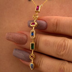 May include: A gold-colored necklace with a series of colorful gemstones. The gemstones include a ruby, a sapphire, an emerald, and other colorful stones. The necklace is suspended from a delicate gold chain.