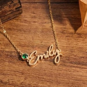 Könnte beinhalten: Eine goldfarbene Halskette mit dem Namen "Emily" in Schreibschrift. Die Kette hat einen kleinen grünen Edelstein und liegt auf einer Holzoberfläche mit getrockneten Blättern.