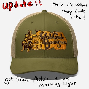 May include: Olive green trucker hat with a tan mesh back. The front features an embroidered design with the text "Hob Gobs and Boomchicks" and a seated figure next to a barrel. The text "update!!" is written above the hat.