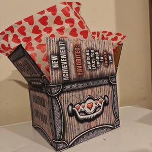 May include: A square cardboard box designed to look like a wooden treasure chest with metal accents. The box features the text NEW ACHIEVEMENT! FAVORITED: Someone Likes You enough to FAVOR you! and PLATINUM LOOT BOX! with red heart tissue paper inside.