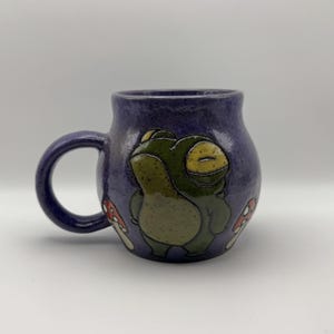 Könnte beinhalten: Ein runder Keramikbecher mit einer glänzenden, violett gesprenkelten Glasur. Auf der Vorderseite befindet sich ein erhabener, handbemalter grüner Frosch mit geschlossenen Augen, flankiert von zwei kleinen rot-weißen Pilzen. Der Becher hat einen Henkel.