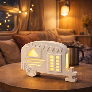 Può includere: Una lampada bianca a forma di roulotte con finestre illuminate e una finestra triangolare. La lampada è su un tavolo rotondo di legno. L'interno della roulotte è illuminato, creando un bagliore caldo. Una tazza in acciaio inossidabile è accanto alla lampada.