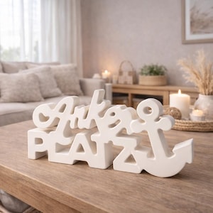 Op de afbeelding: Een witte keramische decoratie op een houten tafel met de tekst "Anker Platz" en een ankersymbool. De woonkamer heeft een beige bank, kaarsen en een ingelijste foto.