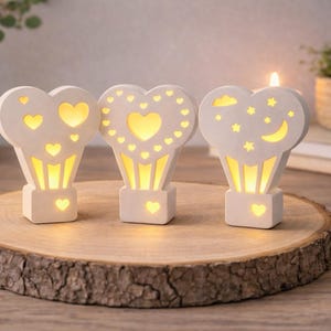 Könnte beinhalten: Drei herzförmige, cremefarbene Dekorationslichter mit ausgeschnittenen Designs, darunter Herzen, Sterne und ein Mond. Die Lichter stehen auf einer Holzscheibe und strahlen warmes Licht aus. Im Hintergrund ist eine brennende Kerze zu sehen.