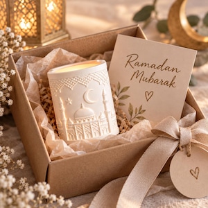 Könnte beinhalten: Eine Geschenkbox mit einem weißen, zylindrischen Kerzenhalter, der ein Reliefdesign einer Moschee, eines Halbmonds und von Sternen aufweist. In der Box befindet sich eine Karte mit dem Text Ramadan Mubarak und einem Herz auf Seidenpapier.