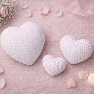 Op de afbeelding: Drie witte hartvormige decoraties met een textuuroppervlak zijn gerangschikt op een roze oppervlak, samen met twee brandende kaarsen, rozenblaadjes en een boeket bloemen. De scène roept een romantisch of feestelijk thema op.