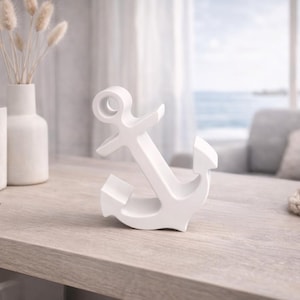 Könnte beinhalten: Ein weißes, ankerförmiges Dekorationsobjekt steht auf einer Holzoberfläche. Der Anker ist der Mittelpunkt, mit einer Vase mit Trockenblumen und Büchern im Hintergrund. Ein nautisch thematisierter Einrichtungsgegenstand.