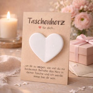Könnte beinhalten: Ein weißes herzförmiges Andenken auf einer Karte mit deutschem Text. Die Karte lautet "Taschenherz" und "für dich..." mit zusätzlichem Text darunter. Eine brennende Kerze, rosa Blumen und eine Geschenkbox befinden sich im Hintergrund.
