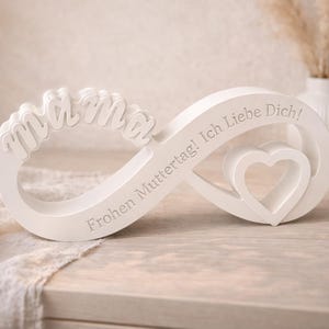 Peut inclure: Un objet décoratif blanc en forme d'infini avec le mot "mama" et les phrases "Frohen Muttertag! Ich Liebe Dich!" gravées. Une forme de cœur est découpée dans le symbole de l'infini. L'objet est sur une surface en bois.