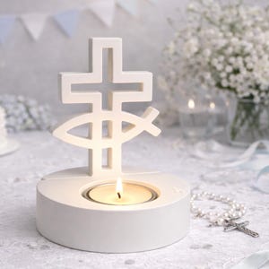 Puede incluir: Un portavelas blanco con un diseño de cruz y pez. El portavelas tiene una base circular con una vela de té encendida en su interior. Un rosario con una cruz se encuentra en el fondo.