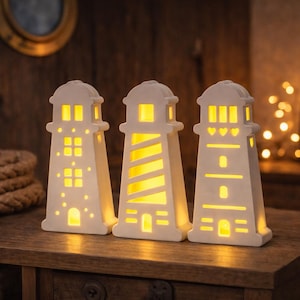 Puede incluir: Tres decoraciones de faro de cerámica blanca con ventanas y diseños iluminados. Los faros están sobre una superficie de madera, con un tema náutico. Las luces dentro de los faros son amarillas.