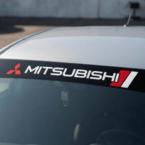Puede incluir: Una franja adhesiva negra aplicada en la parte superior del parabrisas de un coche plateado. El adhesivo presenta el logotipo rojo de tres diamantes de Mitsubishi a la izquierda, el texto blanco MITSUBISHI en el centro y una franja diagonal roja y blanca a la derecha.