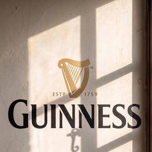 May include: A wall sign featuring the black text GUINNESS and a gold harp logo above the text ESTD 1759. The sign is displayed on a textured off-white surface with a window frame shadow cast across the logo and text, creating a light and dark contrast.