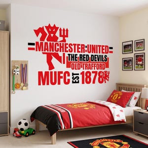 May include: A wall decal featuring the Manchester United logo, the text "MANCHESTER UNITED THE RED DEVILS OLD TRAFFORD MUFC EST 1878", and a red devil graphic. The decal is displayed on a white wall above a bed with red and white bedding.