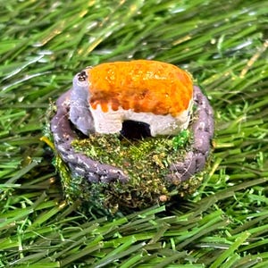 May include: A miniature fairy house with a tan roof and white walls sits atop a mossy, gray stone base. The house is surrounded by green moss and sits on a bed of artificial green grass.