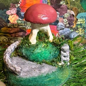 May include: A miniature whimsical mushroom sculpture with a red cap and white stem, perched on a mossy green hill with a stone path. The sculpture is set against a colorful collage background, with a quarter coin for scale.