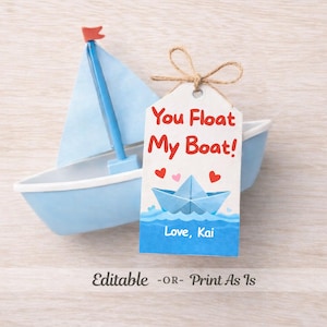以下が含まれることがあります： 白のトリムと青い帆が付いた、水色の玩具のヨット。 「You Float My Boat!」の文字と紙のボートのイラストが描かれた白いタグが、ひもでボートに結び付けられています。 タグには「Kai」という名前も含まれています。
